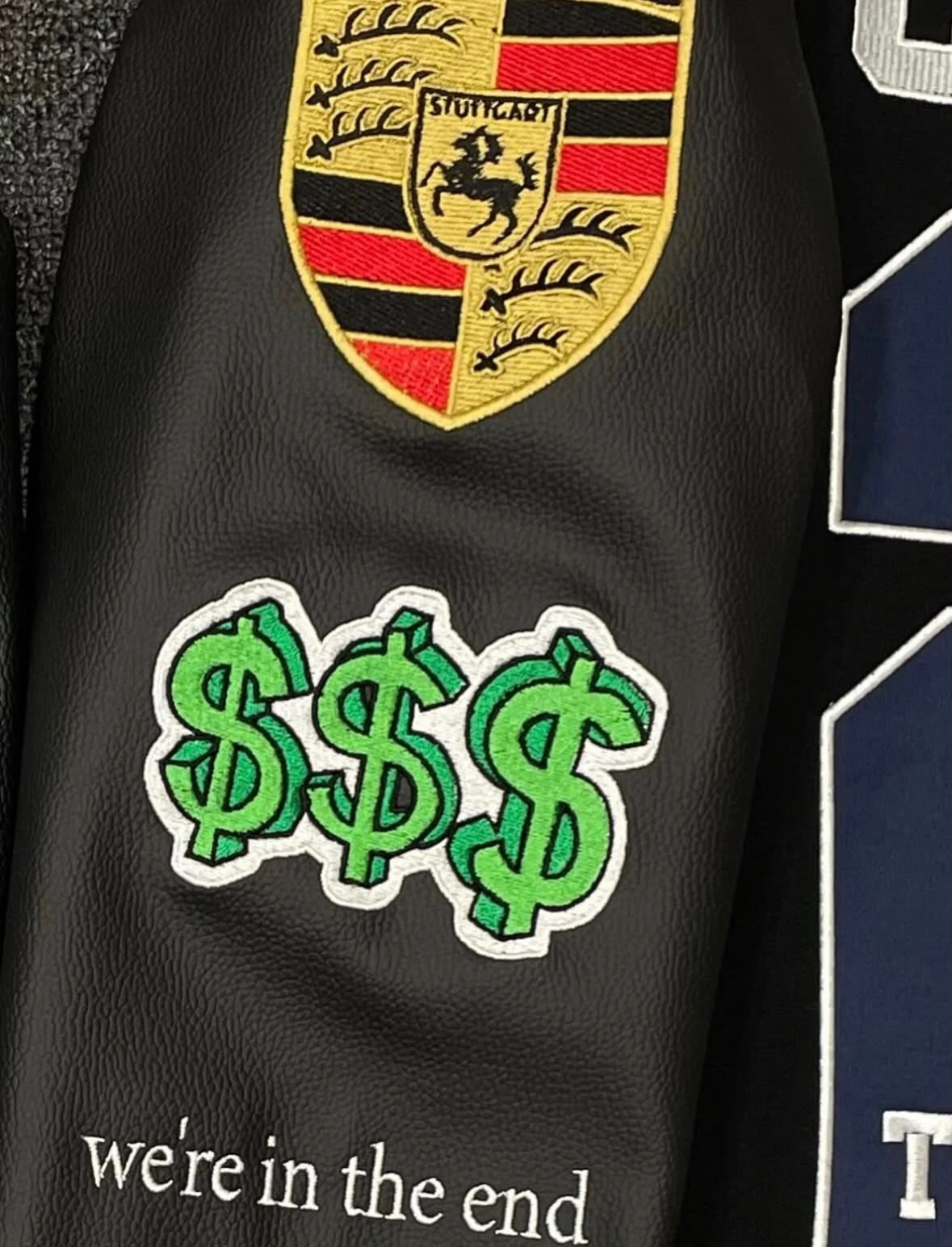 Black leather sleeve with an embroidered crest, a green dollar-sign patch and a script quote